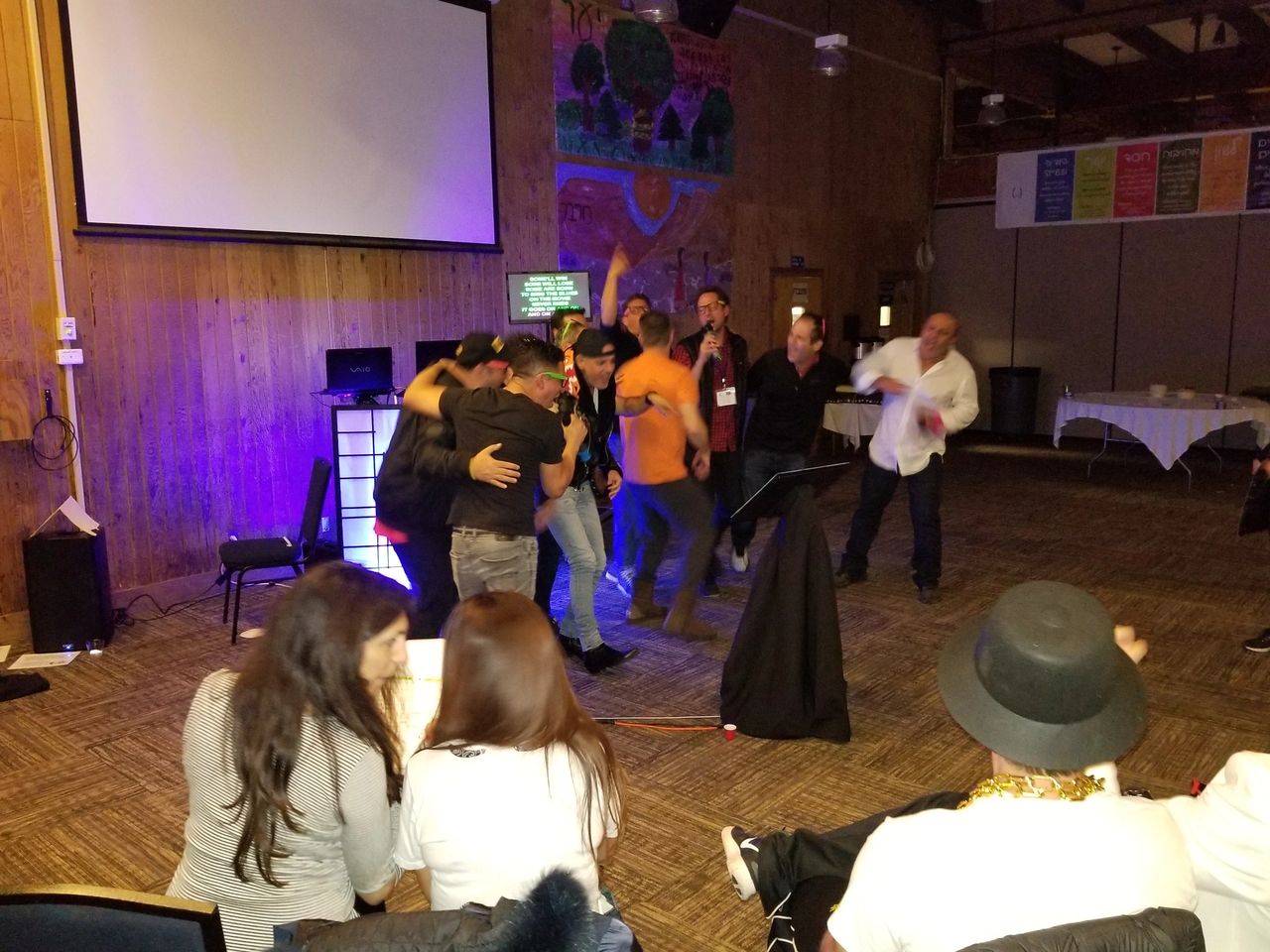 80s theme party. projector screen, some hugging and celebrating, while others sit and watch in a warmly lit room with wood-paneled walls.