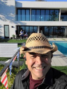 Man wearing a straw hat smiles at the camera in front of a modern house with large windows, a pool, and a person standing by a table in the background.