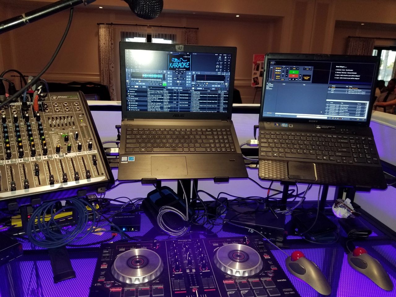 DJ and karaoke setup with a mixer, two laptops displaying DJ and karaoke software, a microphone, cables, and a DJ controller on a lit table.