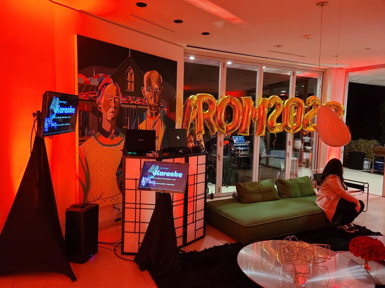 A living room decorated with "PROM 2023" gold balloons, a karaoke setup, colorful lighting, and a mural; a person sits on a green couch.