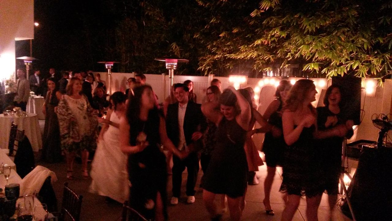 A group of people dance and socialize at an outdoor evening event, with string lights and patio heaters visible in the background.