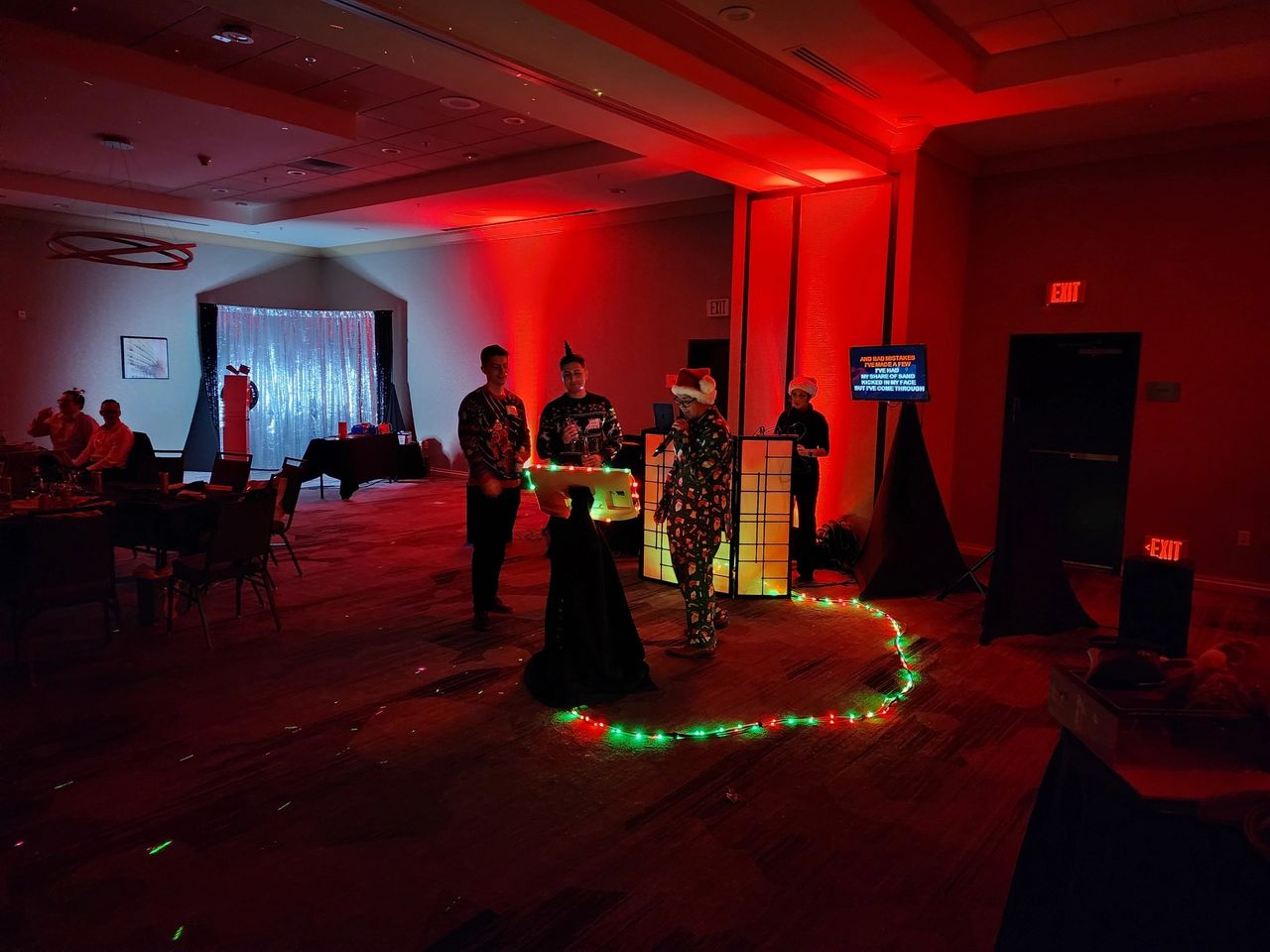 Four people in costumes stand near a lit DJ booth in a dimly lit room with red lighting and string lights on the floor; guests are seated at tables in the background.