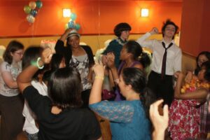 A group of people dance and celebrate together in a decorated room with balloons and orange walls.