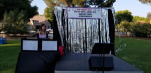 A woman stands behind a DJ booth on an outdoor stage decorated with silver streamers and a sign for a pop-up arts and music fest called “Karaoke Stars #TOPOPP2019.”.