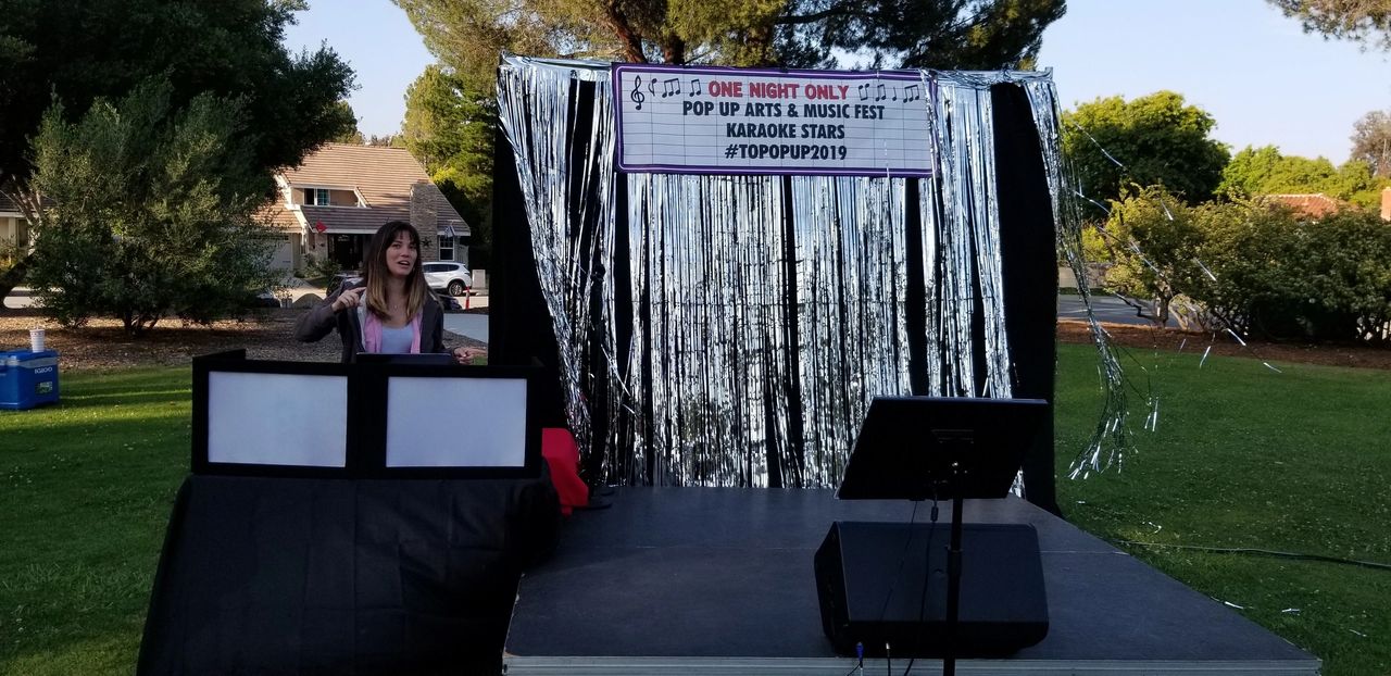 A woman stands behind a DJ booth on an outdoor stage decorated with silver streamers and a sign for a pop-up arts and music fest called “Karaoke Stars #TOPOPP2019.”.
