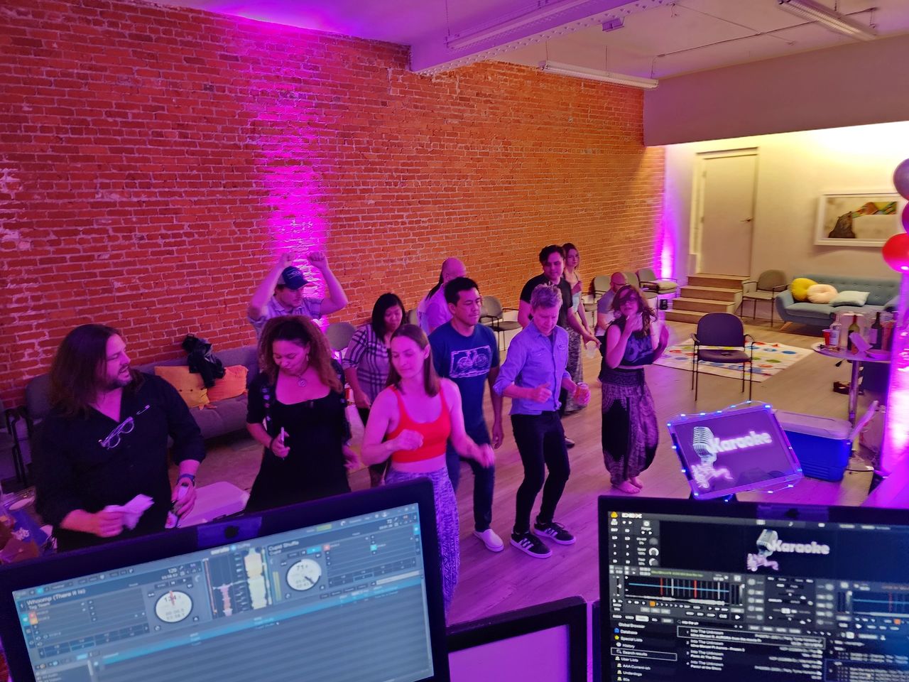 A group of people dance in a room with brick walls and purple lighting, viewed from behind a DJ booth with music equipment and karaoke screens.