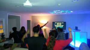 A group of people dance in a living room with dim lighting while karaoke lyrics are displayed on a TV screen in the background.