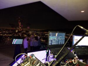 View from a DJ booth overlooking a city at night, with a laptop, cables, and microphones in the foreground and people dancing in the background.