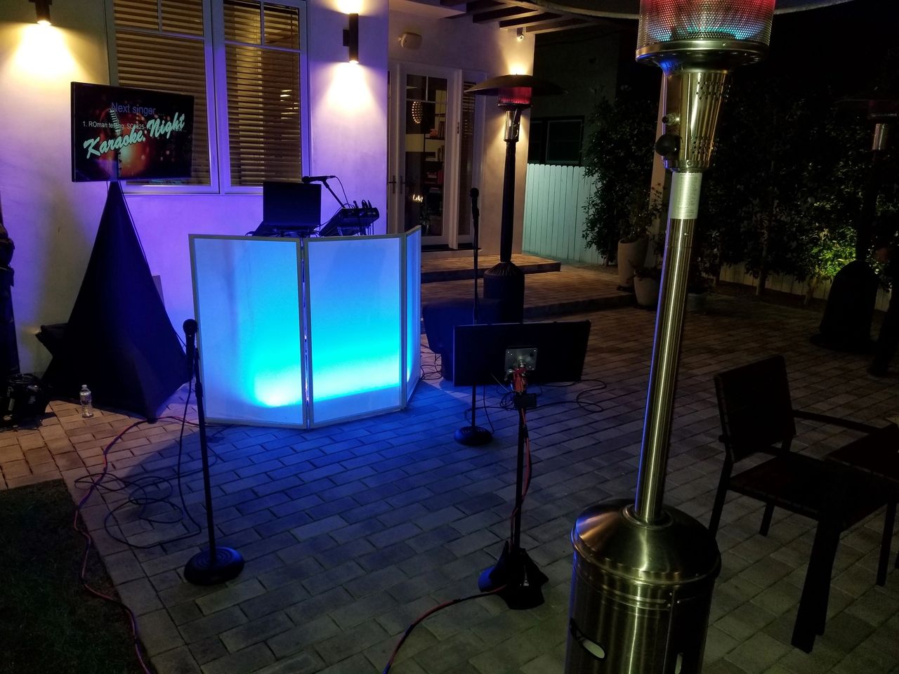 Outdoor karaoke setup with a glowing DJ booth, microphones, speakers, and patio heaters on a tiled patio at night. A "Karaoke Night" sign is visible in the background.