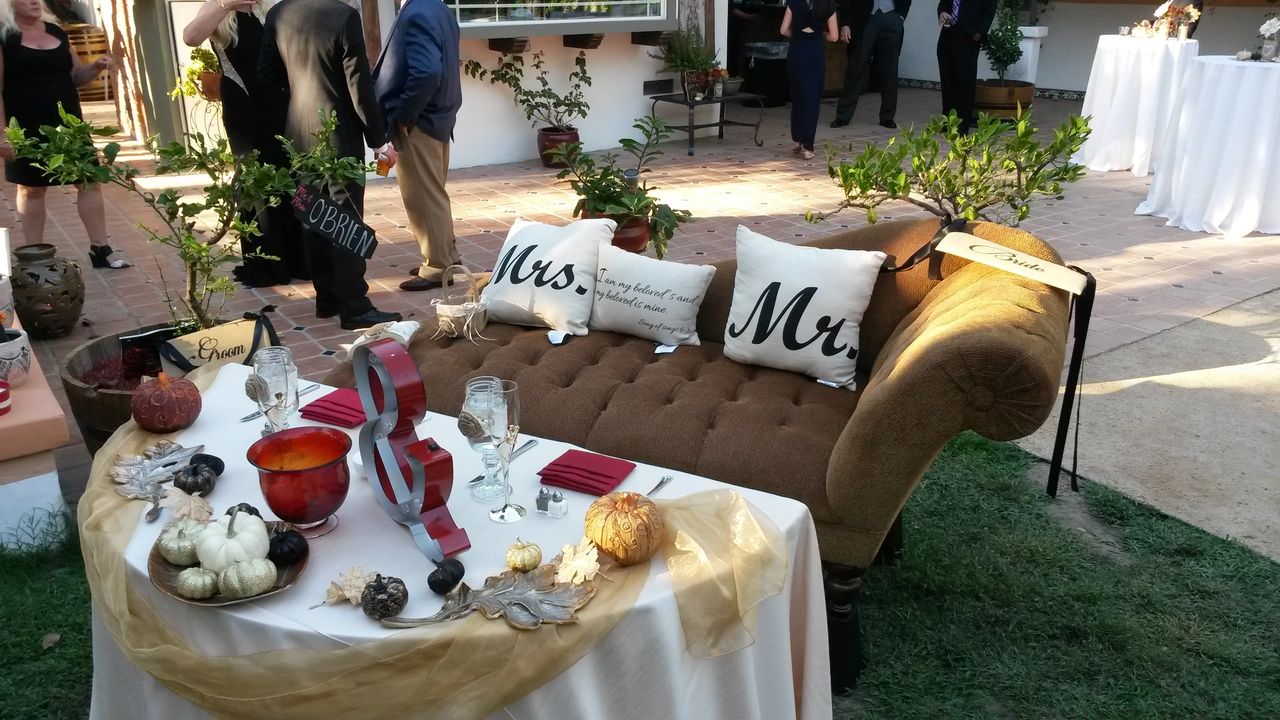 A decorated outdoor wedding table set for two with "Mrs." and "Mr." pillows on a brown loveseat, surrounded by small pumpkins and autumn-themed decor. People are standing in the background.