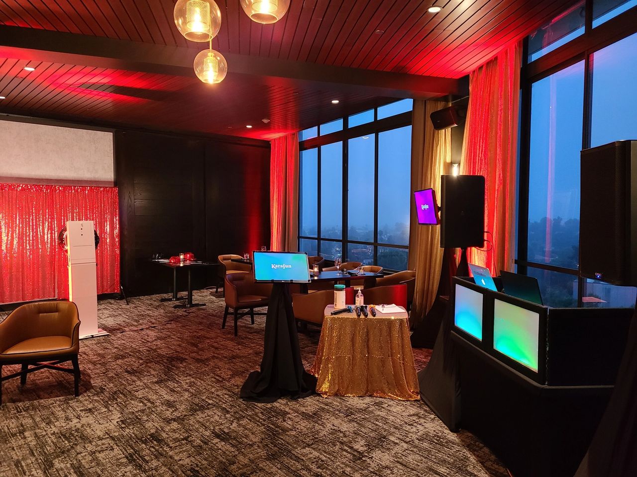 A modern event room with red lighting, lounge chairs, small tables, digital screens, a DJ setup, and large windows showing an evening cityscape.