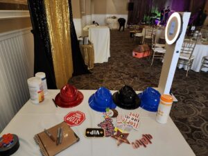 A table with colorful plastic hats, photo booth props, wipes, and a guest book, next to a ring light photo booth at an event venue with empty chairs and tables.