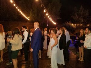 A group of people, some dressed formally, stand and dance under string lights at an outdoor nighttime event.