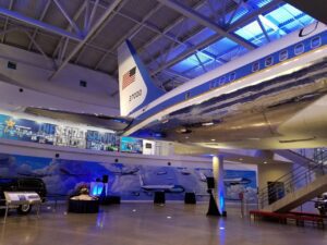 A polished aircraft with tail number 21000 and a U.S. flag is displayed indoors at a museum, with exhibits and murals of planes and Secret Service memorabilia in the background.