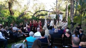 Outdoor wedding ceremony with a bride and groom at the altar, guests seated on chairs, and bridesmaids in red dresses and groomsmen in light suits. Trees and greenery in the background.