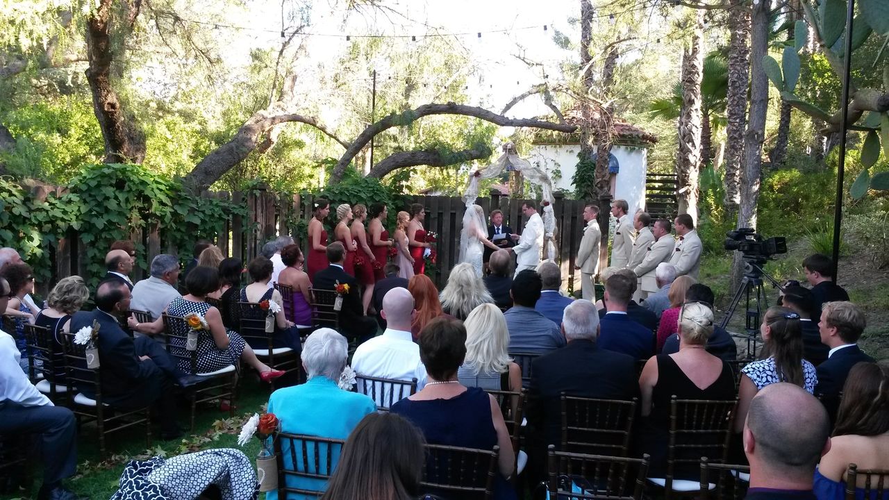 Outdoor wedding ceremony with a bride and groom at the altar, guests seated on chairs, and bridesmaids in red dresses and groomsmen in light suits. Trees and greenery in the background.