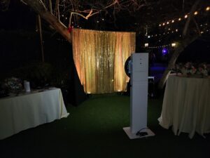 An outdoor photo booth setup with a gold sequin backdrop, a camera stand in front, and two round tables covered with white cloths and floral arrangements. String lights hang above.
