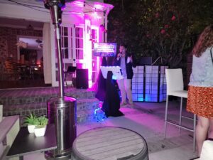 A person sings into a microphone at an outdoor nighttime event with colorful lights, audio equipment, and several people in the background.