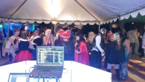 A group of people in costumes are dancing under a tent at a party, viewed from behind a DJ booth with a laptop and equipment.
