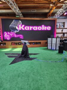 A karaoke setup with a microphone stand and screen on green turf in front of a large display that reads "Karaoke" with musical notes and a microphone graphic.