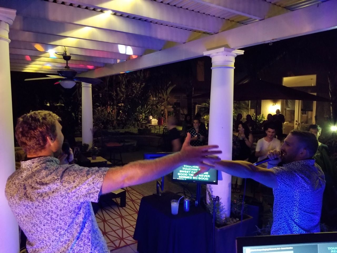 Two men sing karaoke with microphones on an outdoor patio at night, facing each other with arms outstretched, while people watch in the background.