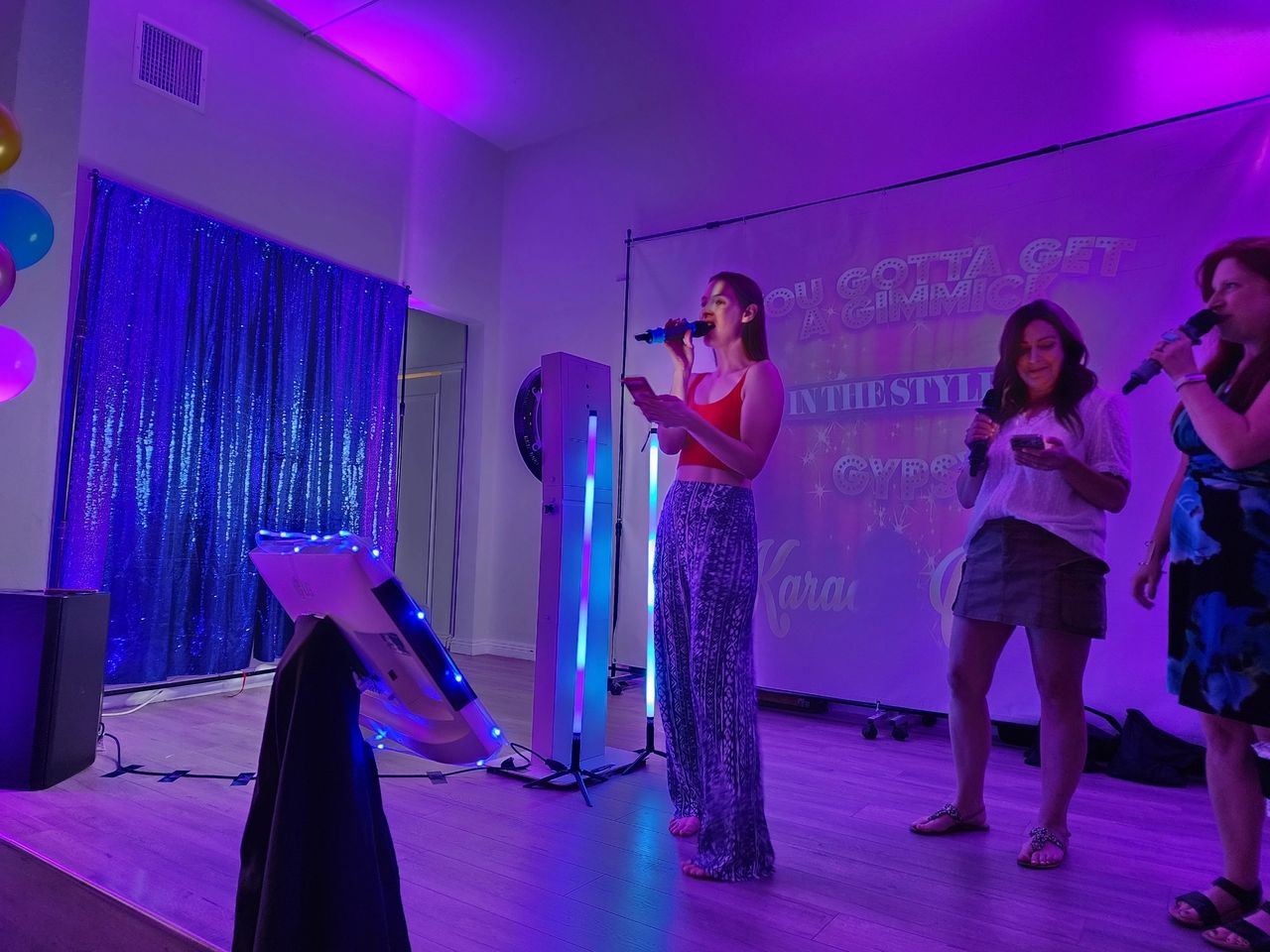 Three women sing karaoke on a small stage with purple lighting, holding microphones. A screen behind them displays song lyrics. Balloons and a sequin curtain are visible to the left.
