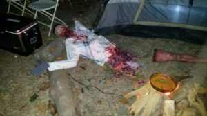 A Halloween prop of a mutilated body with fake blood and entrails is displayed on the ground near a tent and camping supplies.