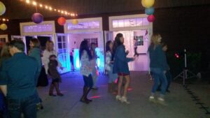 A group of people dance together on a patio at night under string lights and colorful paper lanterns, with a building and open doors in the background.