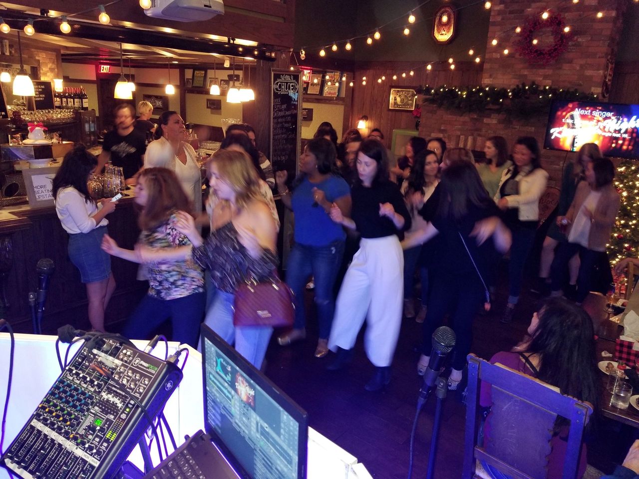 A group of people dance together in a warmly lit bar or restaurant, with music equipment visible in the foreground and festive decorations in the background.